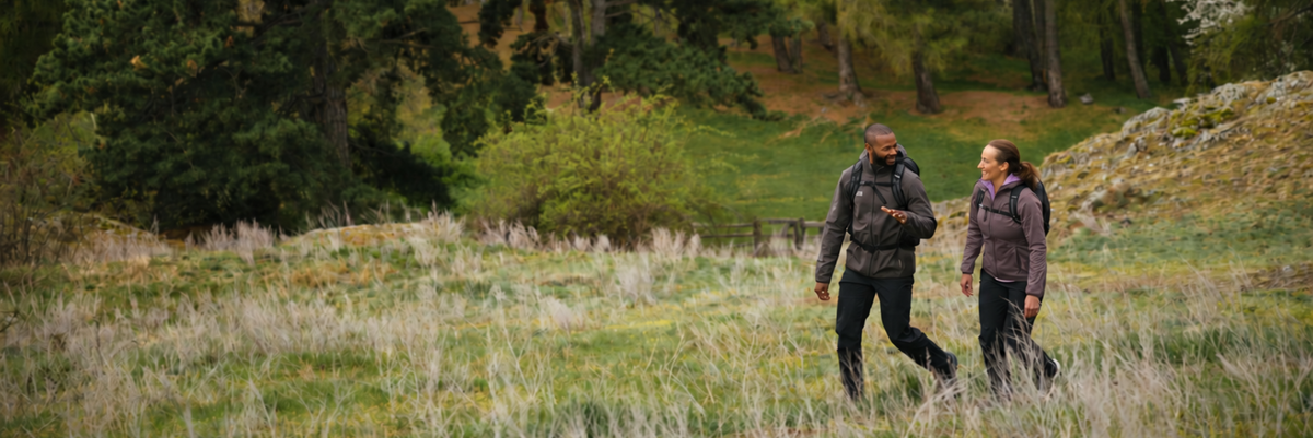 Two people walking through a grassy field with a dense forest in the background, wearing dark outdoor jackets and hiking pants.