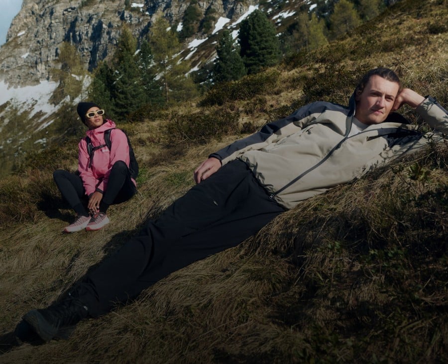 Two out in the country, one sitting and the other lying on the grass, with mountains in the background.
