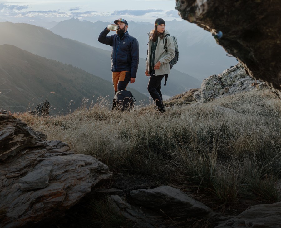 Two out in the country, one sitting and the other lying on the grass, with mountains in the background.